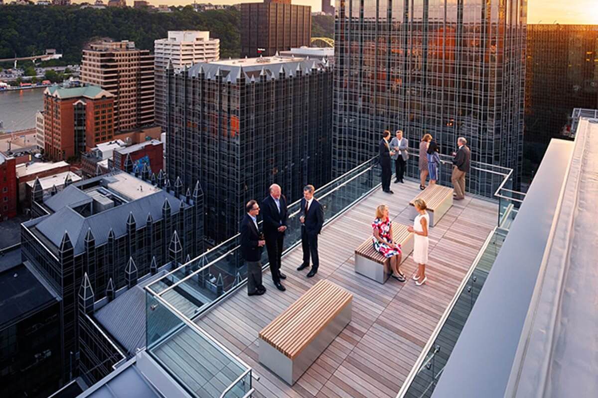 People socialize on a rooftop terrace overlooking a city at sunset.