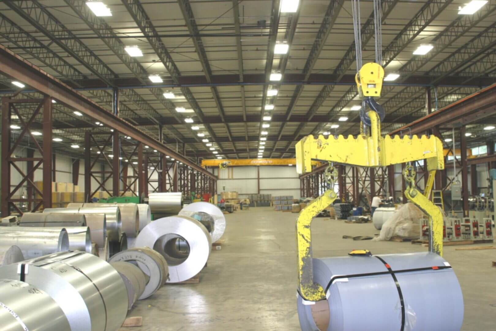 Large rolls of metal sheets stored in a spacious warehouse with overhead cranes.