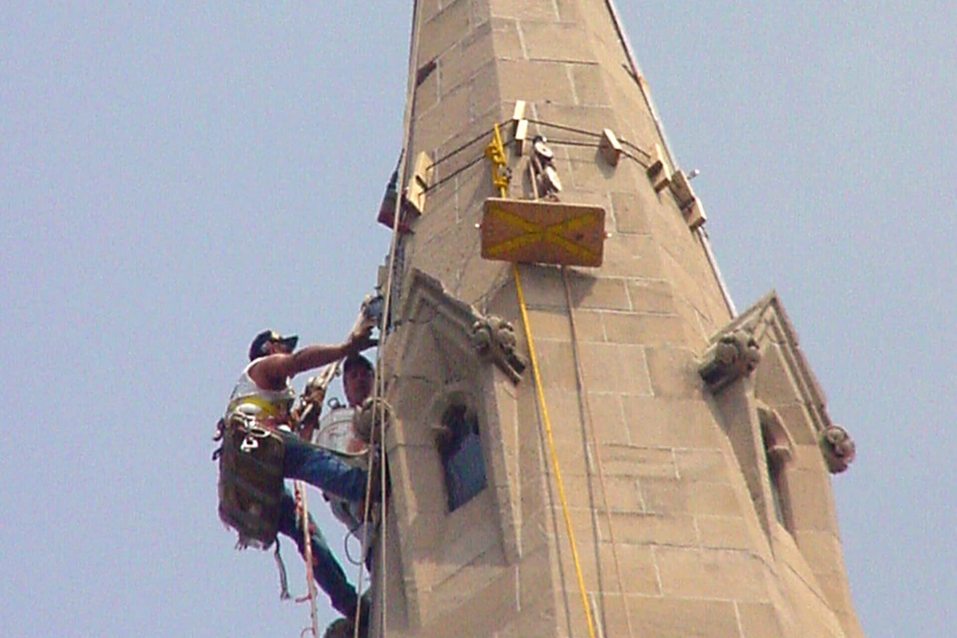 Workers repairing a tall stone tower using ropes and a suspended platform.