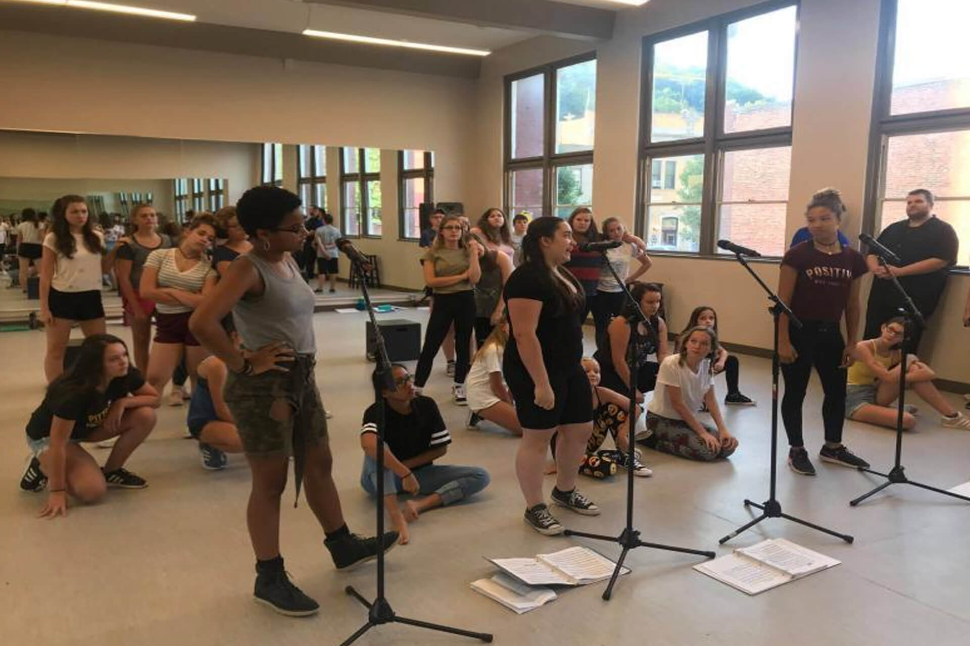 A group of young people rehearsing in a bright room with microphones and papers on the floor.