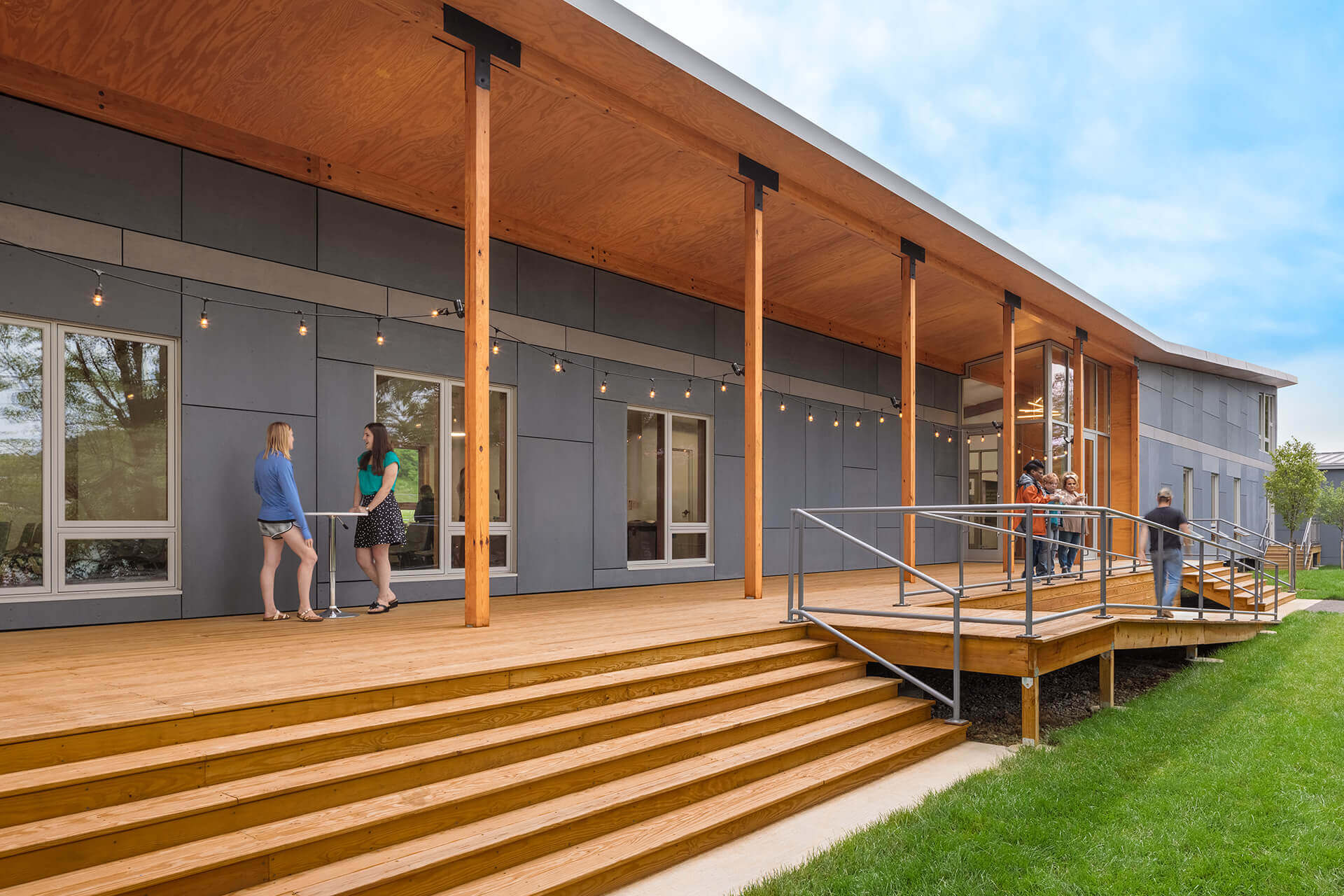 Two people talking on the porch of a modern house with wooden pillars and stairs.