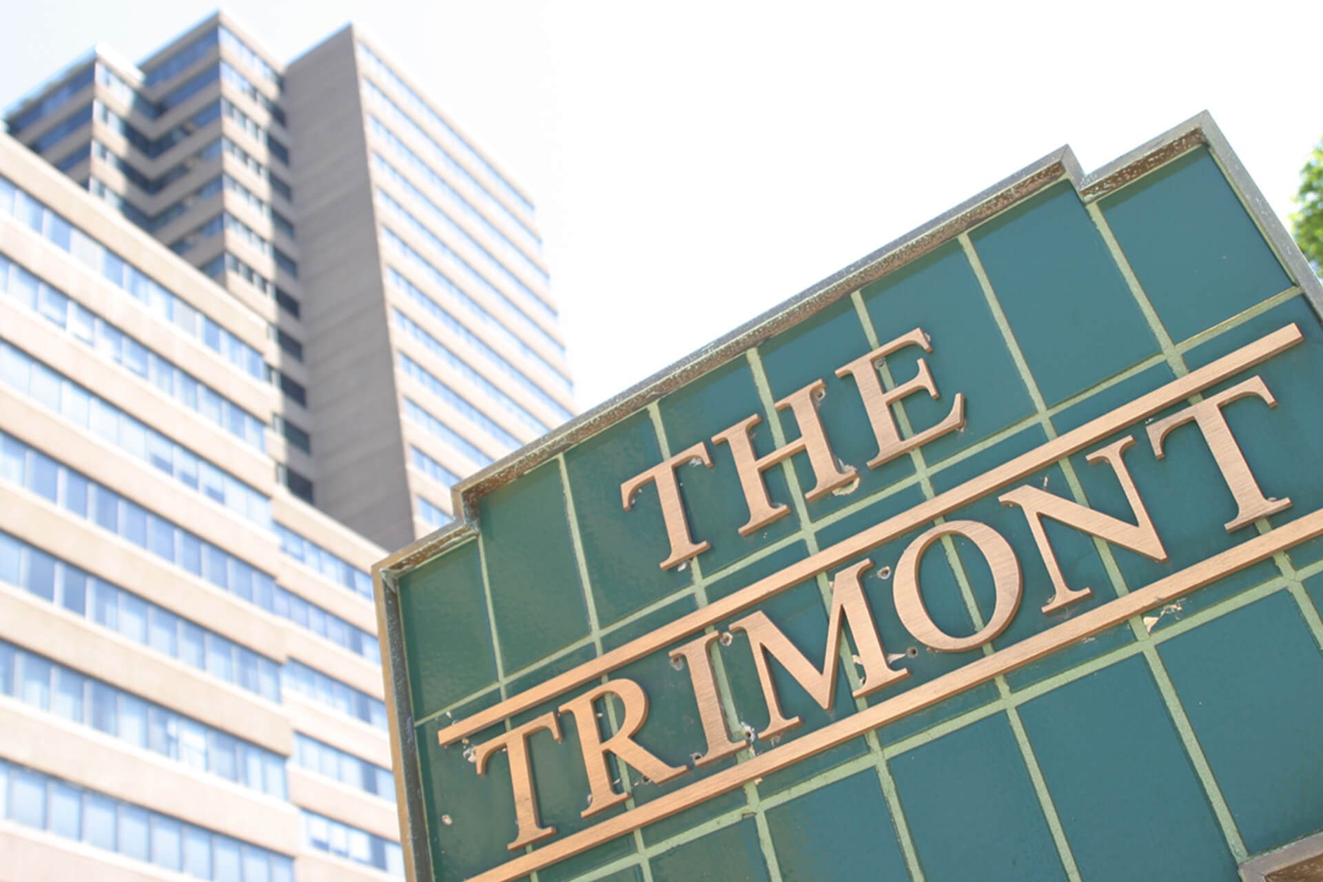 Green and gold sign for The Trimon in front of a modern glass building.